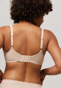 Sostén beige con tirantes ajustables, tejido suave y cierre de gancho y ojo. Presenta una pequeña etiqueta de marca cerca del cierre trasero.