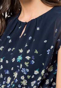 Námořnická modrá blůza s krátkými rukávy, květinový potisk v bílých, zelených a modrých odstínech, má na přední straně nabíraný výstřih a klíčovou dírku.