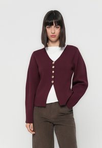 Bordeaux cardigan met een V-halsontwerp, knoopsluiting en lange mouwen. Draag het over een wit shirt, gecombineerd met bruine corduroybroek.