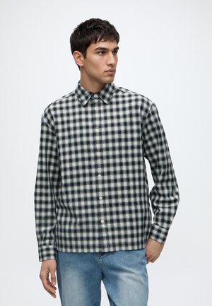Jeune homme portant une chemise à carreaux noir et blanc boutonnée et un jean bleu clair, debout sur un fond blanc.