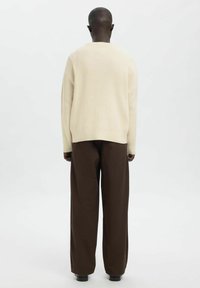 Kreemikas soonikkoes kampsun, mille lõige on lõdvestunud, kombineeritud lahtiste pruuni värvi pükstega, nähtav tagantpoolt, pika varrukaga ja ümmarguse kaelusega.