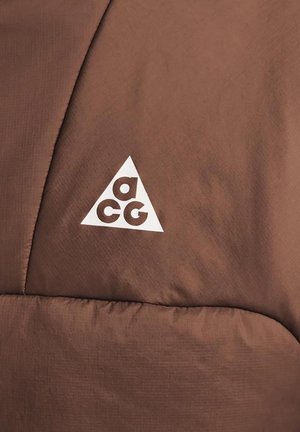 Brun vadderad jacka med en slät textur som har en framträdande vit triangulär logotyp med bokstäverna "a", "c" och "g" inuti.