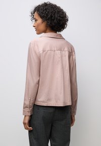 Chemise longue à manches roses claires avec un col, dotée d'une pliure au dos et de poignets boutonnés. Portée avec un pantalon à motifs gris foncé.