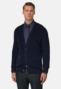Cardigan blu navy in maglia con scollo a V, chiusura con bottoni, due tasche frontali, trama a costine e maniche lunghe indossato sopra una camicia a fantasia.