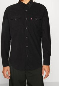 Chemise noire à manches longues avec une patte de boutonnage, dotée de deux poches poitrine, boutons-pression et un empiècement style western.