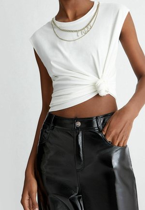 Personne portant un haut blanc sans manches noué à la taille, un pantalon noir brillant et un collier en chaîne argentée superposé.