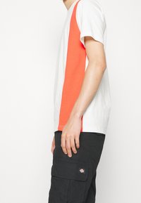 Kortärmad T-shirt med vit kropp och livfull orange sidopanel, gjord av bomull. Kombinerad med svarta cargobyxor med ett märke.