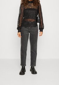 Blouse en dentelle noire à manches longues, col montant et motifs floraux, associée à un jean gris taille haute et des bottines noires.