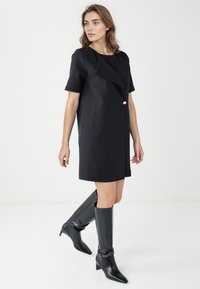 Abito nero a maniche corte con colletto drappeggiato e dettaglio con bottone dorato. Abbinato a stivali in pelle nera al ginocchio con punta affilata.