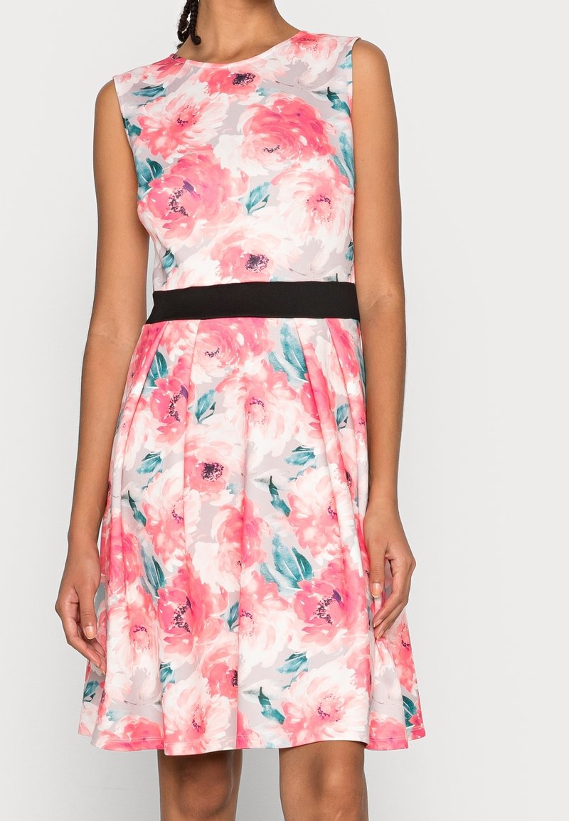 Robe sans manches avec motif floral rose et blanc, feuilles vertes, taille cintrée avec ceinture noire, et jupe plissée se terminant au-dessus des genoux.