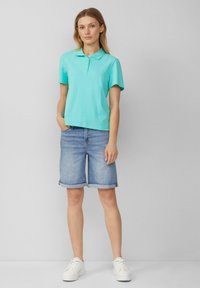 Polo shirt turchese a maniche corte, abbinato a pantaloni corti di jeans risvoltati e sneakers bianche. Il modello sta in piedi con le mani nelle tasche.