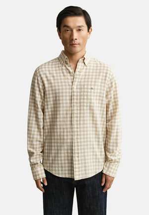 Mann trägt beige-weiß kariertes langärmeliges Hemd mit Knöpfen und Tasche, steht vor weißem Hintergrund.