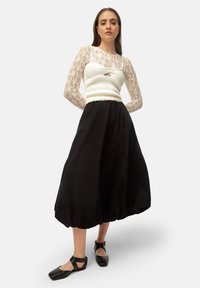 Bluză lungă cu mâneci din dantelă albă, cu un trup ribbed, asortată cu o fustă neagră, voluminoasă. Modelul poartă pantofi negri cu vârf pătrat.