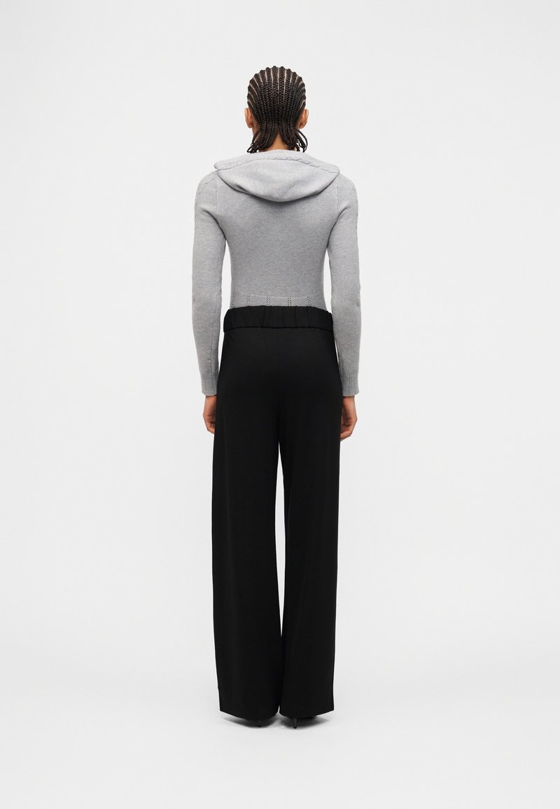 Personne aux cheveux tressés portant un sweat à capuche gris et un pantalon noir à jambes larges, debout de dos sur un fond uni.