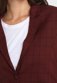 Žena na sobě má bílý tričko s kulatým výstřihem pod burgundským sáčkem s černým kostkovaným vzorem.