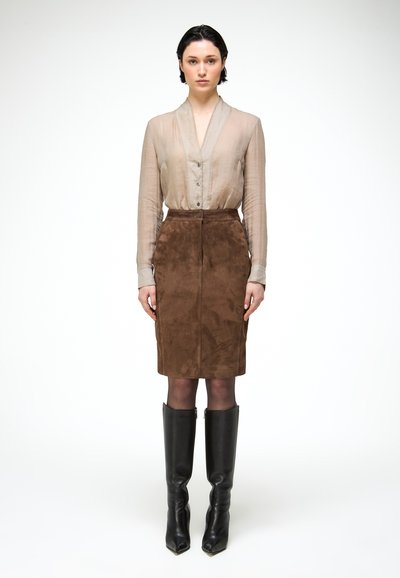 Femme debout portant un chemisier beige à manches longues, une jupe en daim marron longueur genou, des collants noirs et des bottes noires montantes sur un fond blanc.