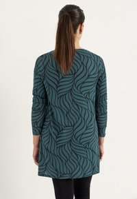 Teal tuniek met lange mouwen, voorzien van een zwart golvenpatroon. Gemaakt van zachte stof, heeft het een losse pasvorm en een ronde zoom. Zwarte leggings worden gedragen.