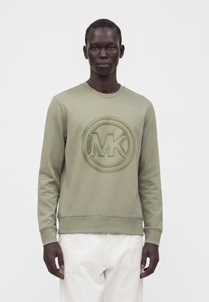 Man draagt licht olijfgroen sweatshirt met geperst rond MK-logo en witte broek tegen een effen achtergrond.