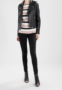 Donna con giacca di pelle nera, top a righe beige-bianco-nero, pantaloni skinny neri e stivaletti nero aperti alla caviglia che si trova davanti a uno sfondo bianco.