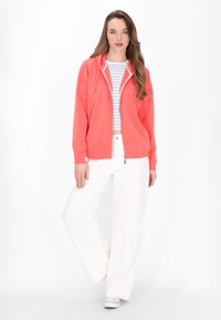 Korallenfarbene Kapuzenjacke mit Reißverschluss, über einem hell gestreiften Hemd getragen. Kombiniert mit weißen weiten Hosen und weißen Sneakers. Glatte Stoffstruktur.