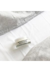 En klar glasburk med nattkräm och ett vitt lock, märkt "Bedtime Beauty Boost." Omgivande tyger är ljusgrå och vita sängkläder.