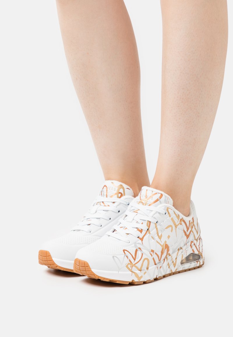 Sneakers bianchi con motivi astratti arancioni, materiale in rete e pelle, punta tonda, suola testurizzata e ammortizzazione visibile nel tallone.