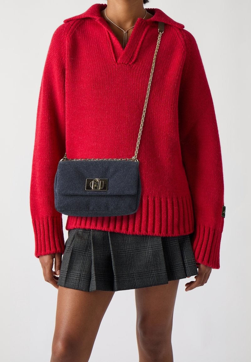 Personne portant un pull en maille rouge, une jupe plissée noire et un petit sac bandoulière bleu foncé avec une chaîne et un fermoir dorés.