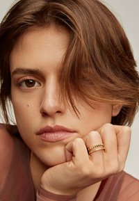 Anillo de banda de oro con pequeños acentos redondos, contra un fondo neutro. Mano descansando sobre la barbilla, mostrando piel suave y un maquillaje sutil.