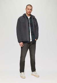 Szary polarowy jacket z kapturem, nałożony na koszulę w kratę. Ciemne dżinsy i jasne sneakersy dopełniają stylizację na jednolitym tle.