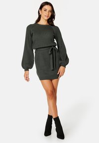 Abito lavorato a maglia di colore verde scuro, caratterizzato da una texture a coste, maniche lunghe a sbuffo, una silhouette aderente e una cintura annodata in vita.