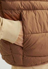 Smeđi puffa prsluk s kvadratnim uzorkom, sa bočnom džepom i bež rebrastim manžetama koje vire ispod.