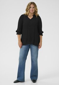 Black V-neck blouse with gathered puff sleeves, paired with light blue flared jeans and black flat shoes. Smooth fabric, relaxed fit.