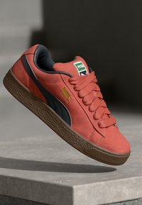 Červené semišové tenisky s čiernymi akcentmi a hnedou gumovou podrážkou. Majú klasický dizajn, hrubé šnúrky a logo Puma na boku.
