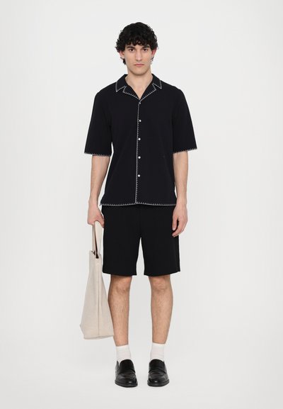 Black short-sleeve button-up shirt with white contrast stitching, paired with black shorts. Model holds a light tan tote bag.