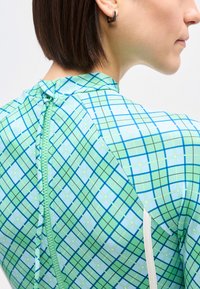 Κοντινό πλάνο ενός ατόμου που φοράει ρούχο με πράσινο και μπλε καρό και φερμουάρ στην πλάτη, δείχνοντας κοντά καστανά μαλλιά και ένα μικρό μαύρο σκουλαρίκι.