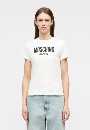 Femme portant un t-shirt blanc Moschino Jeans et un jean bleu clair, debout face à l'avant sur un fond blanc uni.