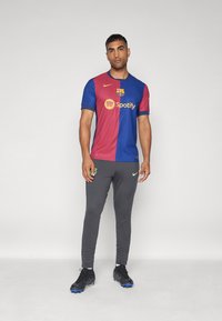 Voetbalshirt van Barcelona met rode en blauwe secties, voorzien van een clublogo en de tekst "Spotify". Grijze sportbroek met groene accenten. Zwarte schoenen.