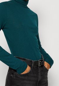 Türkiser Rollkragenpullover aus einem weichen, strukturierten Stoff, kombiniert mit hochgeschnittenen schwarzen Jeans und einem schwarzen Gürtel mit einer silbernen Schnalle.