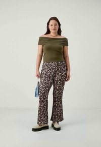 Top off-the-shoulder verde oliva em tecido texturizado, combinado com calças flare de estampa de leopardo. A modelo segura uma pequena bolsa azul e usa sapatos de plataforma bege.