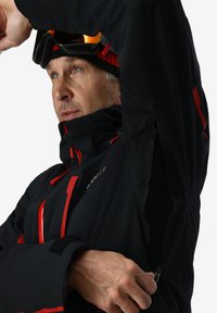 Schwarze Skijacke mit roten Akzenten, hohem Kragen und Reißverschlussbelüftungen unter den Armen. Verfügt über ein elegantes Design und strapazierfähigen Stoff für den Outdoor-Einsatz.