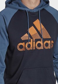 Navy hoodie with blue raglan sleeves, featuring a large orange dotted Adidas logo. Includes drawstring hood and front pocket.