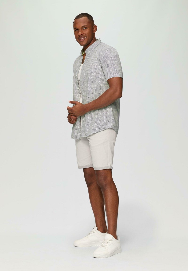 Lichtgewicht korte mouw blouse met patroon in grijs en wit, gecombineerd met beige shorts en witte sneakers,staand op een effen achtergrond.