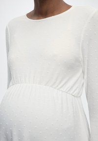 Κοντινό πλάνο ενός ατόμου που φοράει λευκό, μακρυμάνικο φόρεμα εγκυμοσύνης με πουά υφή και ελαστική μέση πάνω από την κοιλιά της εγκυμοσύνης.