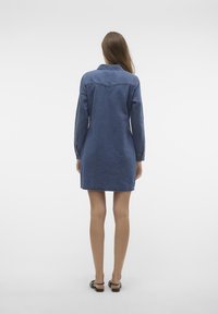 Jeansklänning i mörkblått, långärmad, med krage och detalj i ryggen. Rakt skuren till mitt på låret, med färdig fåll, bärs med sandaler.