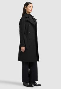Donna in piedi di lato che indossa un trench coat doppiopetto nero, dolcevita nero, pantaloni a gamba larga e stivali neri con tacco.