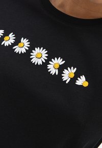 Zwarte katoenen t-shirt met een rij witte madeliefjes met gele centra. Het ontwerp is in de stof geborduurd.