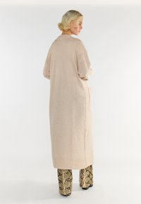 Blondyna stojąca bokiem, ubrana w długi beżowy kardigan o teksturowym wykończeniu z dużymi kieszeniami oraz spodnie w wzór węża, na tle białym.