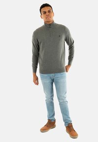 Maglione grigio con colletto a zip a un quarto, tessuto strutturato, indossato sopra jeans blu chiaro e stivali color beige con finitura liscia.