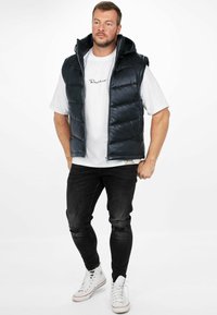 Gilet imbottito nero con cappuccio, indossato sopra una camicia bianca. Presenta una chiusura a zip e tasche frontali. Abbinato a jeans neri strappati e scarpe da ginnastica bianche.