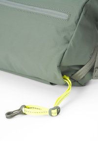 Grüner Stoff-Rucksack mit strukturiertem Oberflächen, ausgestattet mit einer grauen Reißverschlusstasche und einem leuchtend gelben Tragegurt mit einem Plastikclip zur Befestigung.